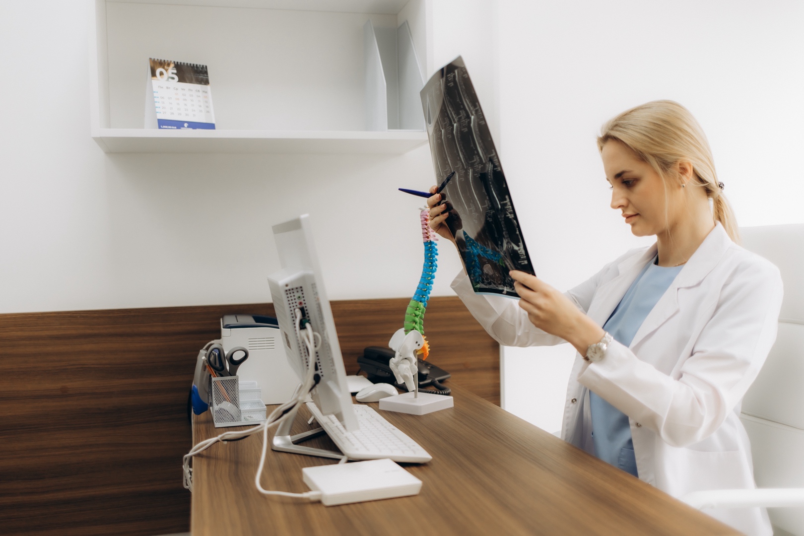 A female doctor with blonde hair is sitting at a wooden desk, examining an X-ray. She wears a white coat and holds a pen. A model of a spine and a computer are on the desk. A calendar appears on a shelf in the background. The setting is a medical office.